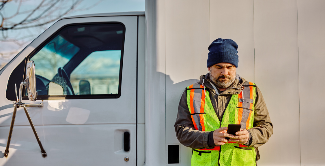 A truck driver in a safety vest and beanie checks his phone while leaning against the side of his white truck.