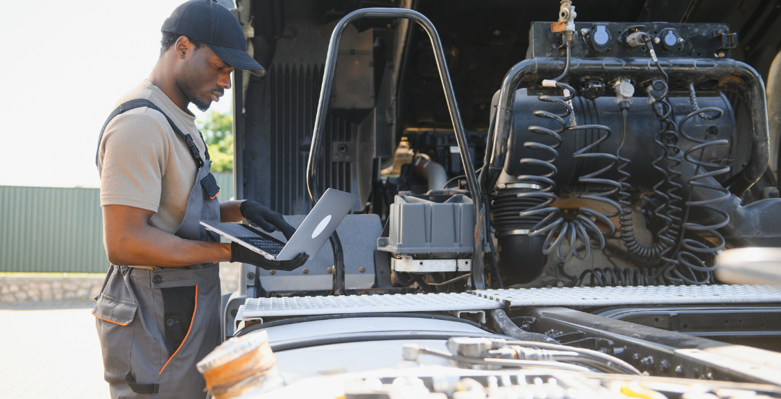 A mechanic or technician uses a laptop to diagnose or work on a truck engine. The person is wearing work clothes and gloves and is focused on the screen.