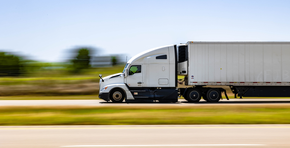 a semi truck moving along a rural road with some trees in the background