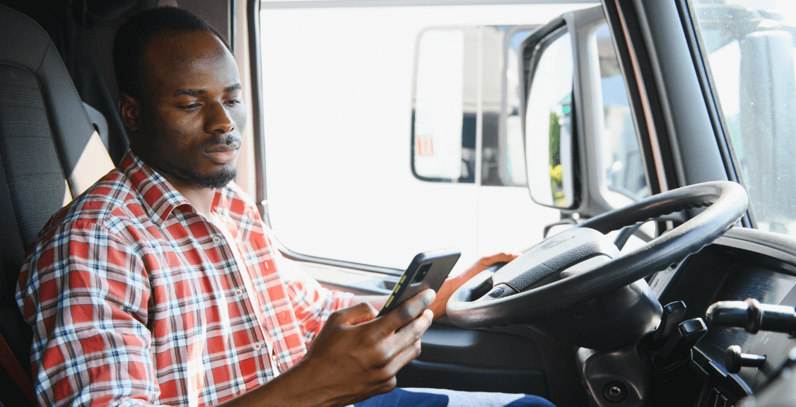 A truck driver is looking at his smartphone while seated in the driver's seat of his commercial vehicle.